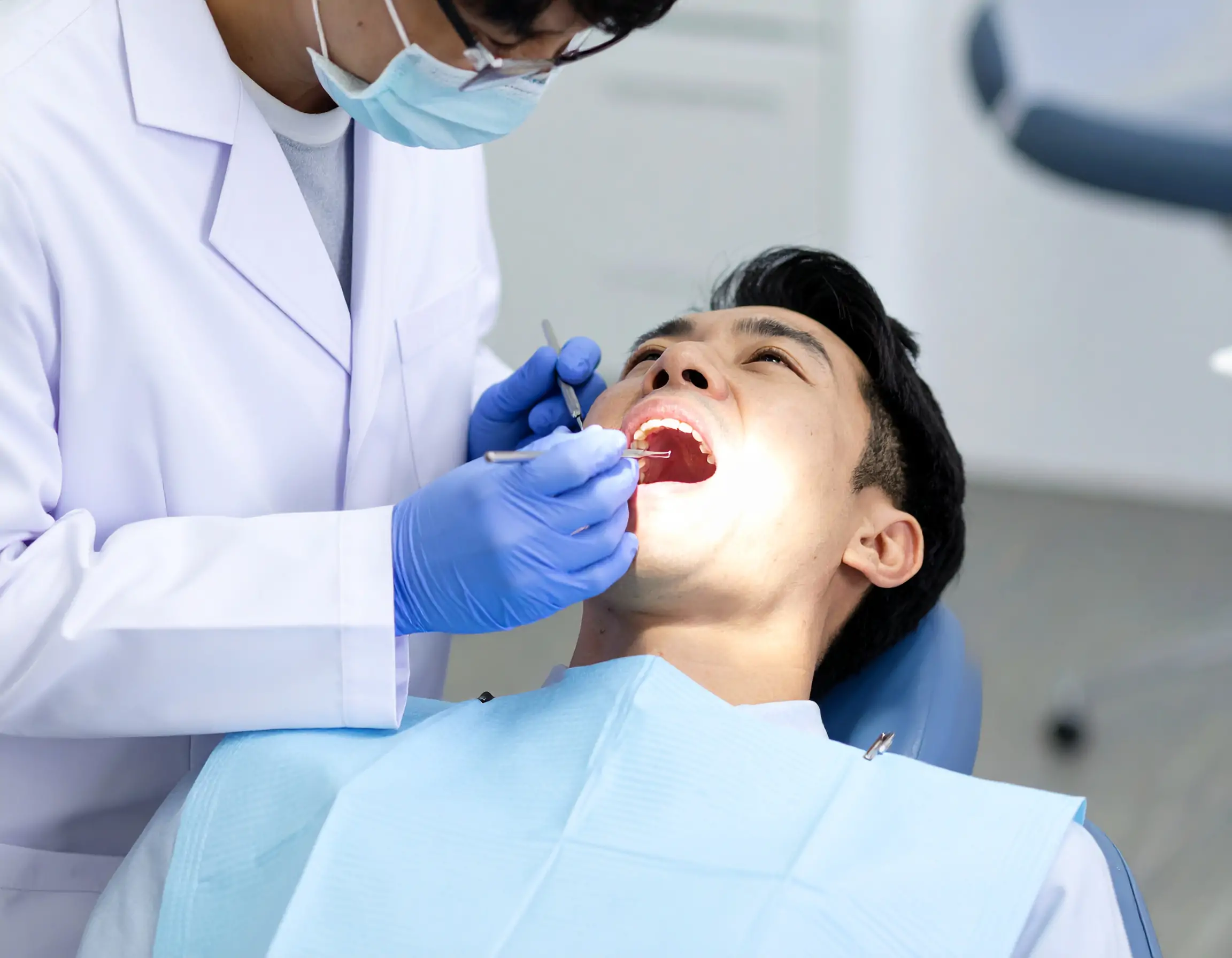 歯科用椅子に患者のよだれかけと彼の隣に座っている歯科医を持つ若い男。彼は歯科用顕微鏡を使用して自分の歯を見て、歯科用バーと鏡を持っています。